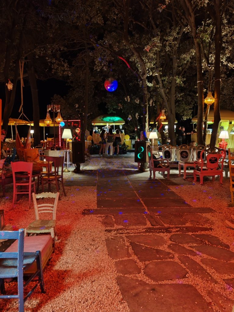 Un locale all'aperto illuminato da luci colorate, con tavoli e sedie disposti lungo un vialetto. Alcune persone sono in gruppo. Un grande disco appeso ad un albero riflette luci colorate nel buio della sera.